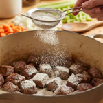 Une main saupoudre de la farine à l'aide d'une passoire sur des morceaux de viande dorée dans une cocotte. Légumes hachés en arrière-plan.